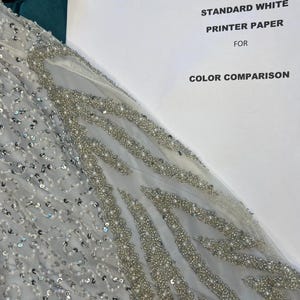 May include: A detailed view of a light grey fabric adorned with sequins and beads, creating elaborate patterns. The image includes a sheet of paper with the text "STANDARD WHITE PRINTER PAPER FOR COLOR COMPARISON" for colour reference.