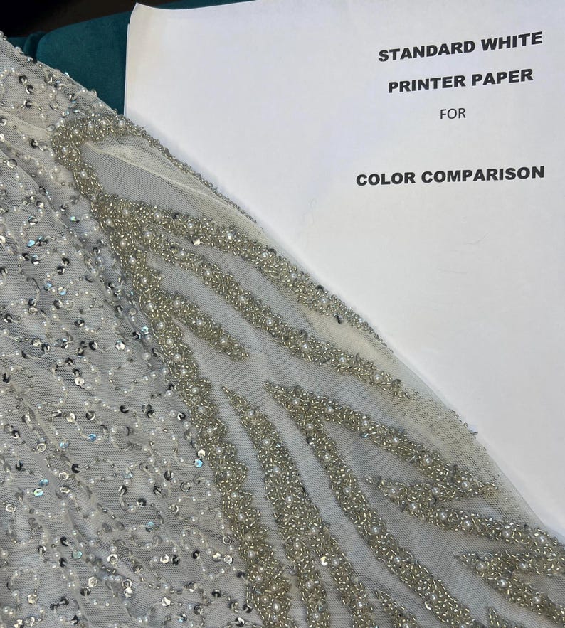 May include: A detailed view of a light grey fabric adorned with sequins and beads, creating elaborate patterns. The image includes a sheet of paper with the text "STANDARD WHITE PRINTER PAPER FOR COLOR COMPARISON" for colour reference.