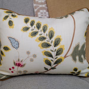 May include: Rectangular decorative pillow with a cream background and a botanical pattern featuring yellow, green, and pink floral elements. The pillow has a brown trim and is placed on a gray surface.