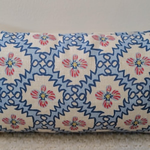 May include: A rectangular throw pillow featuring an off-white base fabric with a repeating pattern of blue zigzag diamond shapes. Inside each diamond is a small floral motif with pink petals and a yellow center, creating a symmetrical design.