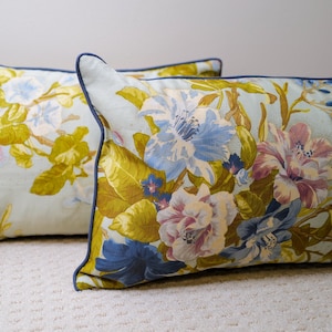 May include: Two rectangular throw pillows with floral patterns. The pillows feature a light blue background with large, colorful flowers in shades of pink, blue, and yellow. The edges are trimmed with a dark blue piping.