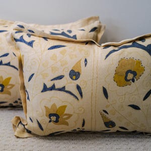 May include: Two decorative rectangular pillows with a cream-colored background and a repeating pattern of blue and yellow floral and leaf designs. The pillows have ruffled edges.