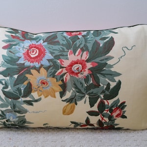 May include: A rectangular throw pillow with an off-white background featuring a botanical print of passion flowers in shades of blue, yellow, and pink, surrounded by dark green foliage and thin, winding tendrils, finished with dark green piping.