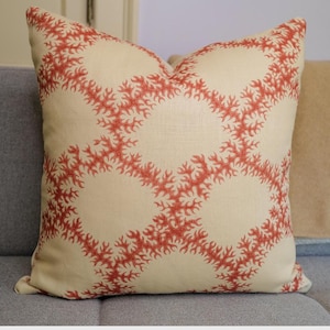 May include: Square decorative pillow with a cream-colored background and a repeating coral-red pattern. The pillow is placed on a gray sofa, and the design adds a touch of coastal style to home decor.