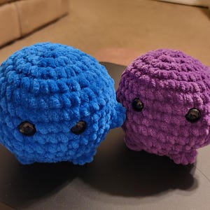 Peut inclure: Deux créatures rondes en peluche crochetée avec du fil doux et texturé. L'une est bleu vif et l'autre est violette. Toutes deux présentent de petits yeux de sécurité noirs et brillants ainsi que de courts appendices trapus.