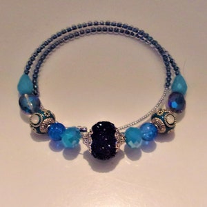 May include: A blue beaded bracelet with a central dark blue stone. The bracelet features various shades of blue beads, including turquoise and dark blue, and is designed with a spiral shape. The bracelet is a piece of jewellery.