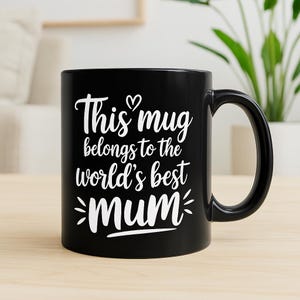 Puede incluir: Una taza de cerámica negra con una inscripción blanca que dice "This mug belongs to the world's best mum". La taza tiene un asa curva y está sobre una superficie clara.