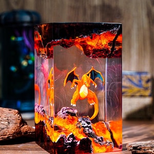 May include: A clear resin cube featuring an orange and yellow Charizard figure set against a fiery, lava-like background. The cube has a rectangular shape and is displayed on a wooden surface.