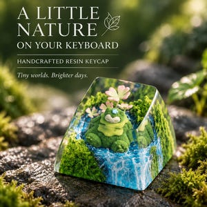 Può includere: Un tasto in resina trasparente con una creatura verde in miniatura vicino a una cascata blu e fiori rosa. Il testo recita: A LITTLE NATURE ON YOUR KEYBOARD, HANDCRAFTED RESIN KEYCAP, Tiny worlds. Brighter days. Il design include texture muschiate.