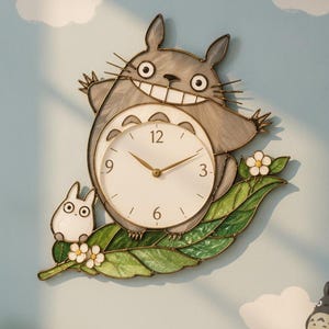 May include: A wall clock shaped like a large grey creature with a wide smile, featuring a white circular clock face with gold hands and numbers 3, 6, 9, and 12. The creature sits on a large green leaf with small white flowers and a tiny white figure.