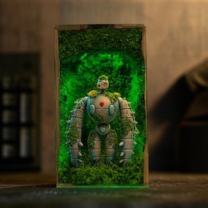 May include: A rectangular resin block containing a miniature robot figure covered in green moss and small yellow flowers. The robot has a light blue, segmented body with a red heart on its chest and a glowing green light illuminating the mossy interior.