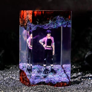 May include: A rectangular resin block containing a miniature figure of a person with short dark hair, wearing a pink jacket, black crop top, and black trousers. The base and top of the block feature textured, iridescent purple and dark red material.