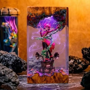 以下が含まれることがあります： ピンクの髪と赤と黒の衣装を着たフィギュアが特徴の装飾的な樹脂ブロック。 フィギュアは、ホーンスピーカーを備えたヴィンテージラジオの上に立っています。 樹脂ブロックは木製のベースと紫と緑のデザインです。