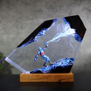 May include: A resin sculpture of Captain America with a hammer, set within a clear, angled block. The sculpture has blue lightning effects and is mounted on a wooden base. The design incorporates dark blue accents, creating a unique superhero-themed artwork.
