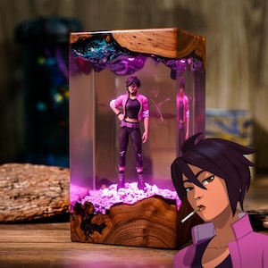 Puede incluir: Una pieza de arte de resina rectangular con base y tapa de madera. En el interior, una figura de una mujer con chaqueta rosa y pantalón negro se encuentra sobre un lecho de piedras moradas. La resina es transparente con detalles morados y azules.