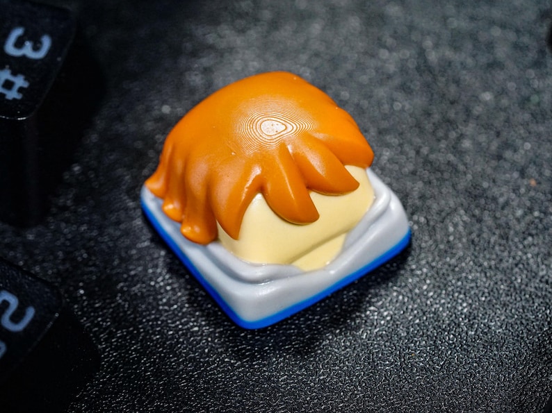 Peut inclure: Un capuchon de touche personnalis&eacute; avec un design brun, orange et blanc. Le capuchon de touche a une base blanche avec une bordure bleue et un design brun, orange et blanc sur le dessus. Le design ressemble aux cheveux d'une personne.