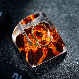 Könnte beinhalten: Ein klarer, abgewinkelter Keycap mit einem feurigen, lavaartigen Design in Rot-, Orange- und Gelbtönen. Ein goldfarbener Ring ist in das Harz eingebettet. Der Keycap befindet sich auf einer dunklen, strukturierten Oberfläche, mit Tastaturtasten sichtbar.