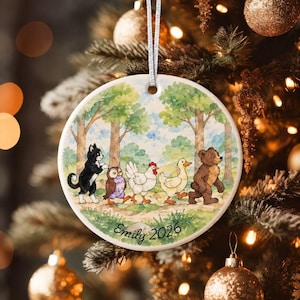 Op de afbeelding: Een rond wit keramieken ornament met een aquarelillustratie van een zwart-witte kat, een uil, een witte kip, een gele eend en een bruine beer die in een rij door een bos lopen. De tekst Emily 2026 staat onderaan op het ornament gedrukt.