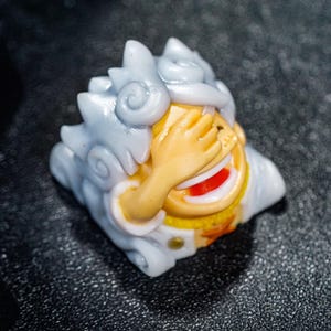 Peut inclure: Un capuchon de touche en r&eacute;sine blanc et jaune avec un visage de dessin anim&eacute;. Le visage se couvre les yeux d'une main et a une bouche grande ouverte avec une langue rouge.