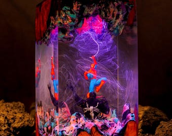 Könnte beinhalten: Eine rechteckige Harzlampe mit einer Miniaturfigur von Spider-Man, die über einer schwarzen Venom-Figur schwebt. Das klare Harz enthält wirbelnde violette und blaue Raucheffekte, ergänzt durch einen Sockel und eine Oberseite aus Holz.