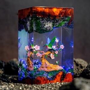 May include: A rectangular resin block lamp featuring a miniature scene of a green hummingbird perched on a flowering tree branch with pink blossoms. The base and top consist of textured, dark reddish-brown wood with green mossy accents.