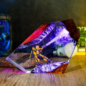 May include: A yellow and black action figure is encased in a clear, geometric resin block. The figure is positioned on a rocky surface, with a purple energy beam emanating from its weapon. The resin block has a wood-grain base and a red and purple top.