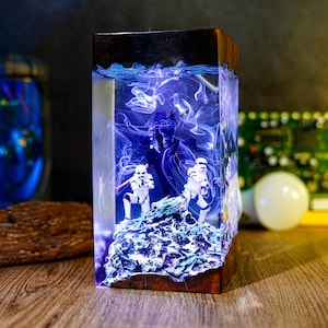 Könnte beinhalten: Ein rechteckiges Kunstwerk aus Harz mit Star Wars-Motiv. Es zeigt Sturmtruppen auf einer felsigen Oberfläche, eine dunkle Figur und blaue und weiße Wirbeleffekte in klarem Harz. Die Basis und die Oberseite bestehen aus dunklem Holz.