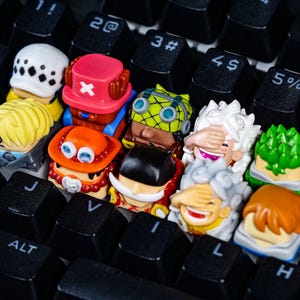 Peut inclure: Un ensemble de dix touches de clavier color&eacute;es repr&eacute;sentant des personnages de dessins anim&eacute;s avec diff&eacute;rentes expressions faciales. Les touches sont dispos&eacute;es sur un clavier noir.