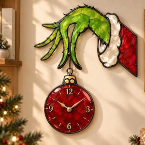 May include: A stained glass wall clock featuring a green hand with long fingers wearing a white and red cuff, holding a circular red ornament clock face with gold numbers 3, 6, 9, and 12 and gold clock hands, all outlined in black metal.