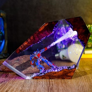 May include: A resin sculpture featuring a blue and red robot figure, set within a clear, angled block. The sculpture has a wood-like base with purple and white accents, and a bright light effect.