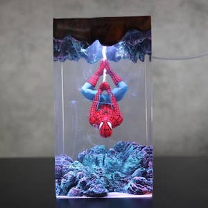 May include: A rectangular resin art piece featuring a Spiderman figure hanging upside down. The figure is red and blue, suspended above blue and green coral-like formations. The top has a brown and blue design.