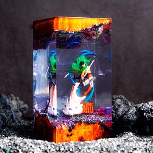 May include: A rectangular resin block featuring a miniature anime character in a green cape and brown gear, suspended within clear material. The top and bottom sections consist of textured, multi-colored wood and resin with orange and dark tones.