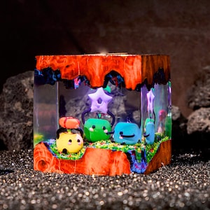 May include: A square resin block featuring a natural wood frame with a vibrant orange and red grain. Inside the clear resin, three small, colorful cartoon-like figures are displayed on a green base, including a yellow, green, and blue character.