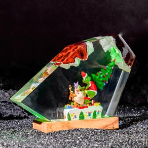 Könnte beinhalten: Eine Skulptur aus klarem Kunstharz mit einer grünen Figur im roten Anzug, die einen grünen Weihnachtsbaum und einen Sack hält, begleitet von einem braunen Hund. Die Szene steht auf einem weißen Sockel mit bunten Geschenkmustern auf Holz.
