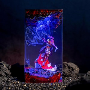 May include: A rectangular resin block containing a miniature anime figure in a dynamic pose. The figure wears a brown jacket and red scarf, standing on a textured base. Swirling white smoke-like patterns are suspended in the blue-lit resin.