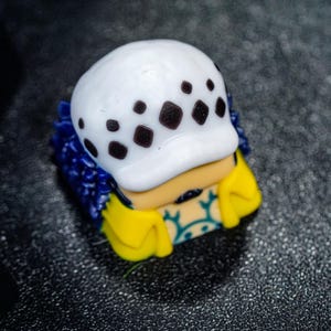 Peut inclure: Un capuchon de clavier blanc et bleu avec un motif de cr&acirc;ne noir et blanc. Le capuchon de clavier est en forme de t&ecirc;te avec un chapeau et une veste jaune et bleue.
