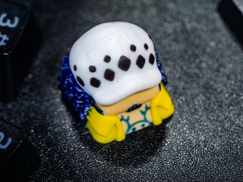 Peut inclure: Un capuchon de clavier blanc et bleu avec un motif de cr&acirc;ne noir et blanc. Le capuchon de clavier est en forme de t&ecirc;te avec un chapeau et une veste jaune et bleue.