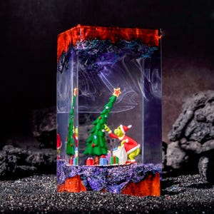 Könnte beinhalten: Ein rechteckiger Harzblock mit einer grünen Figur im roten Weihnachtsmannkostüm, die einen grünen Weihnachtsbaum mit einem gelben Stern schmückt. Die Basis und die Oberseite sind mit violetten und orangen Tönen texturiert, während weiße Wirbel im Harz.