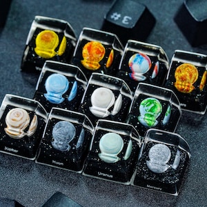 Puede incluir: Un conjunto de 10 teclas con diferentes planetas de nuestro sistema solar. Cada tecla está hecha de plástico transparente con una base negra y una imagen colorida de un planeta en su interior. Las teclas están dispuestas en una fila, con los planetas en orden desde el sol. Las teclas están etiquetadas con los nombres de los planetas: Mercurio, Venus, Tierra, Marte, Júpiter, Saturno, Urano, Neptuno, Plutón y el Sol.