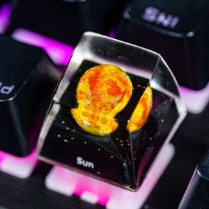 May include: A clear resin keycap with a depiction of the sun in orange and yellow hues. The keycap is labeled "Sun" and is sitting on a black keyboard with pink lighting.