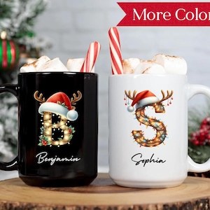 May include: Two ceramic mugs, one black and one white, filled with marshmallows and candy canes. The black mug features a letter "B" decorated with Christmas lights and a Santa hat, with the name "Benjamin" below. The white mug has a letter "S" with similar decorations and the name "Sophia".