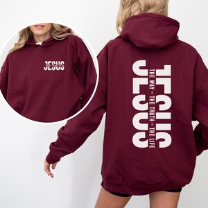 May include: A maroon hoodie featuring the word "JESUS" in white, printed vertically on the back, with the text "THE WAY - THE TRUTH - THE LIFE." The front of the hoodie also displays the word "JESUS" in white. The hoodie has a hood and long sleeves.