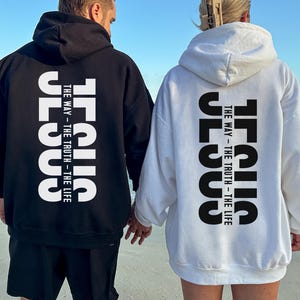 May include: Black and white hoodies featuring the word "JESUS" printed vertically on the back, with the text "THE WAY - THE TRUTH - THE LIFE." Two people are seen wearing the hoodies while standing on a beach.