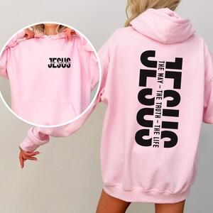 May include: A pale pink hoodie featuring the word "JESUS" in black on the front and the words "JESUS THE WAY - THE TRUTH - THE LIFE" printed vertically on the back. The hoodie has a hood and a front pocket.