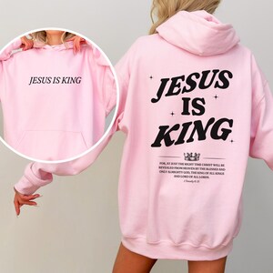 May include: Light pink hoodie with the text "JESUS IS KING" on the front and back. The back of the hoodie features the words "JESUS IS KING" in a bold, black font, along with a bible verse. The hoodie has a hood and a front pocket.