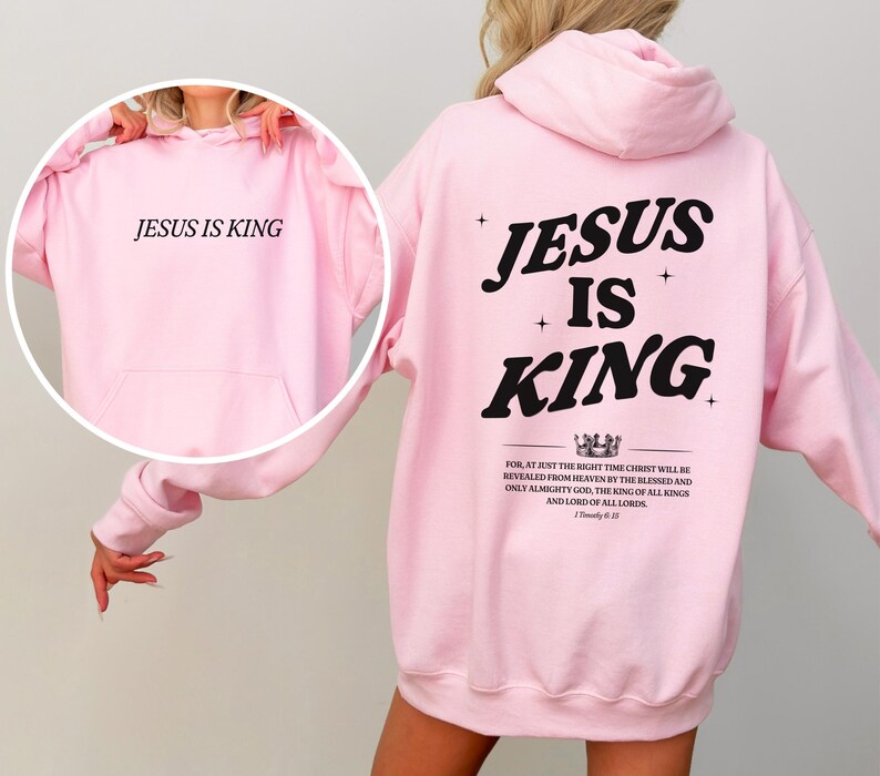 May include: Light pink hoodie with the text "JESUS IS KING" on the front and back. The back of the hoodie features the words "JESUS IS KING" in a bold, black font, along with a bible verse. The hoodie has a hood and a front pocket.