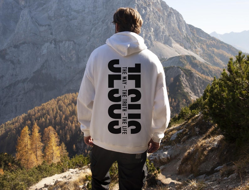 May include: A white hoodie featuring the word "JESUS" printed vertically in black on the back, with the text "THE WAY - THE TRUTH - THE LIFE". The person is on a mountain path with a scenic backdrop.