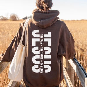May include: A brown hoodie with the word "JESUS" printed vertically in white on the back, with the text "THE WAY - THE TRUTH - THE LIFE". The person is wearing a white tote bag and standing on a wooden bridge.