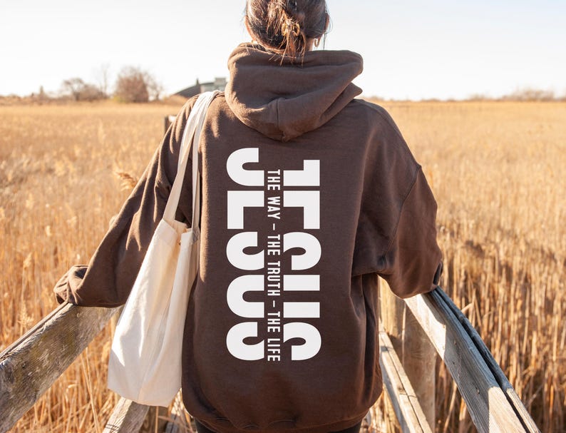 May include: A brown hoodie with the word "JESUS" printed vertically in white on the back, with the text "THE WAY - THE TRUTH - THE LIFE". The person is wearing a white tote bag and standing on a wooden bridge.