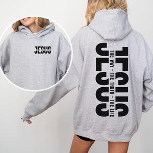 May include: A light grey hoodie with the word "JESUS" printed in black on the front and back. The back of the hoodie also features the text "THE WAY - THE TRUTH - THE LIFE" in black. The hoodie has a hood and long sleeves.